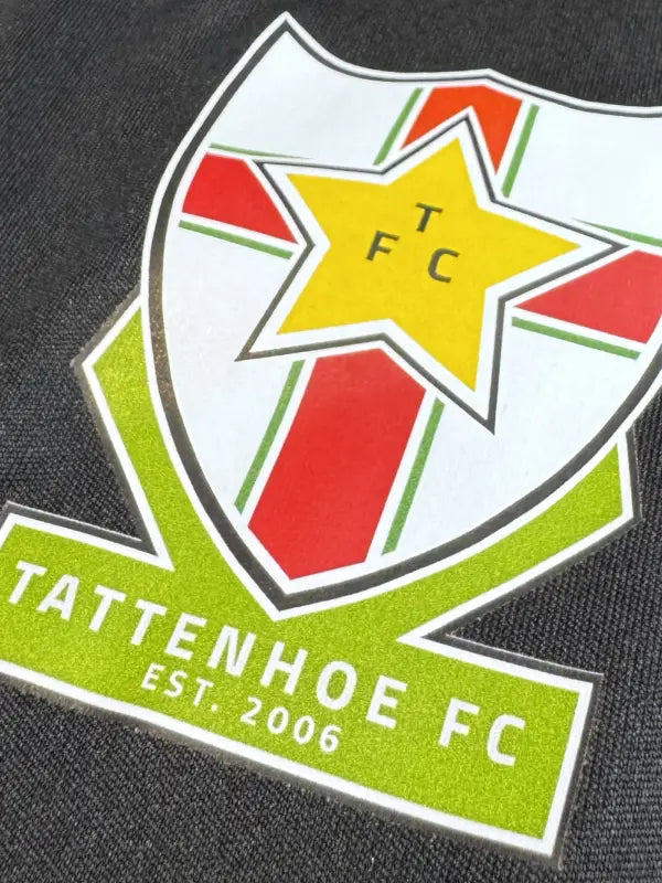 Close-up of a detailed football club crest iron on transfer on a dark fabric, featuring a white shield with a yellow star, red and green cross details.