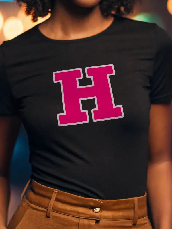 A black woman wearing a black t-shirt with a bold pink iron on initials transfer letter H, bordered in white, styled in college block font.