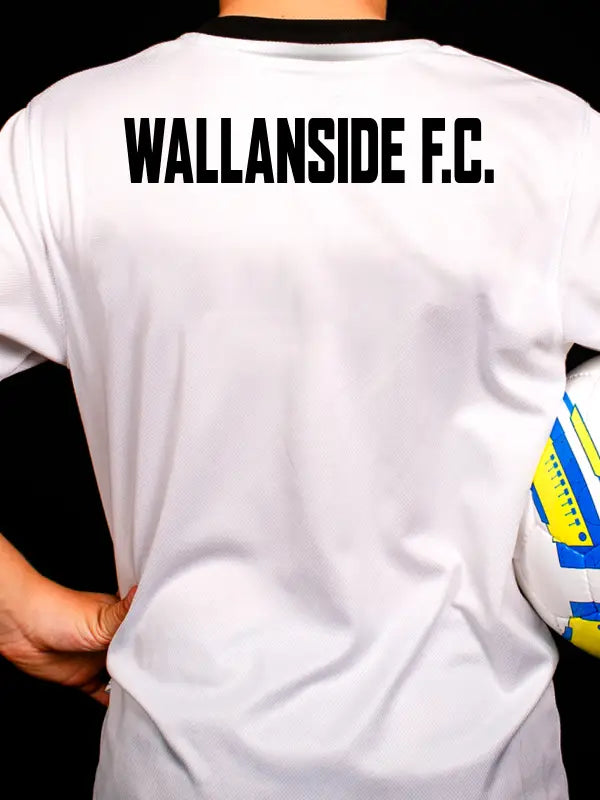 Back view of white football shirt with bold black WALLANSIDE F.C. iron on team name applied across the shoulders, player holding a football against a dark background