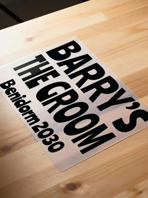 Iron on stag party transfer on clear carrier film laid on a wooden surface, showing bold black text reading “Nobby’s Stag Fest Benidorm 2030”.