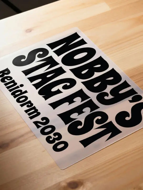 Iron on stag party transfer on clear carrier film laid on a wooden surface, showing bold black text reading “Nobby’s Stag Fest Benidorm 2030”.